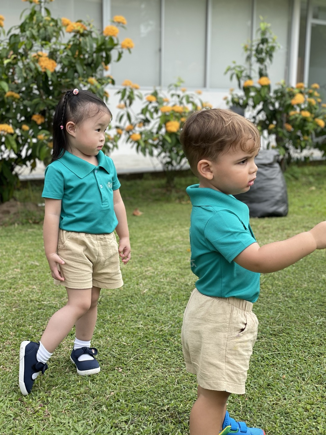 Two toddlers in Little Forest polo shirts standing near each other on the grass, each looking in a different direction
