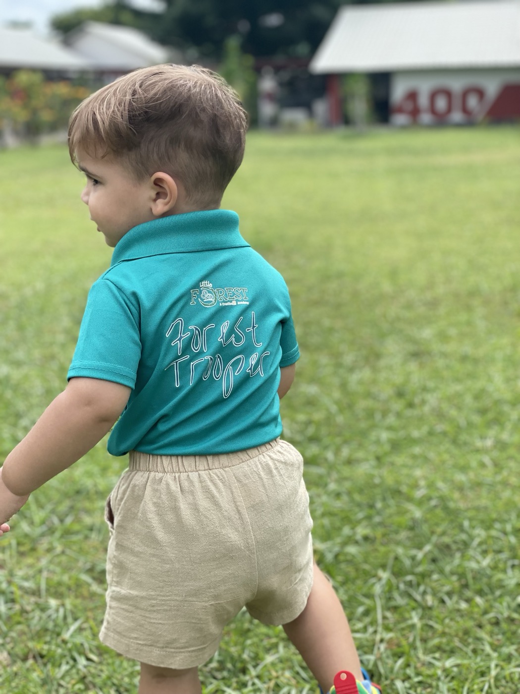 A toddler in a Little Forest polo shirt walking across the grass, glancing off to one side