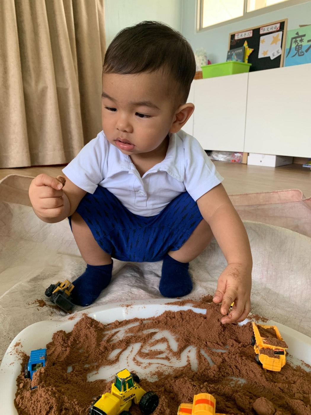 A toddler crouched at a tray of cocoa powder and toy diggers at Little Forest, pinching the powder between his fingers