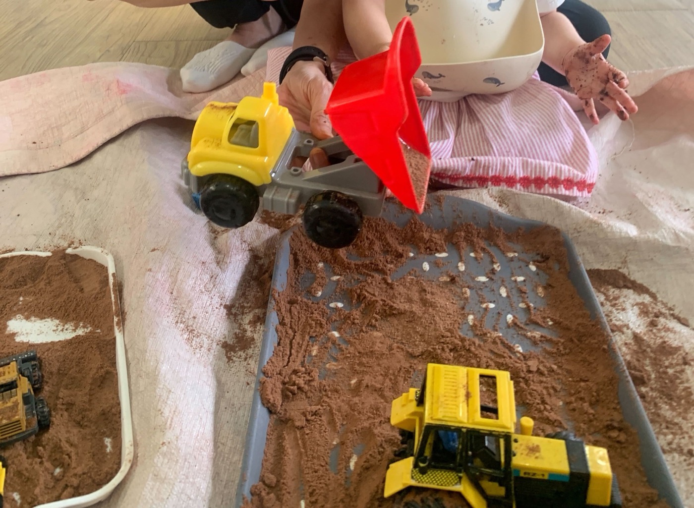 A toddler's hand, covered in cocoa powder, reaching over a tray of cocoa and toy diggers at a Little Forest class