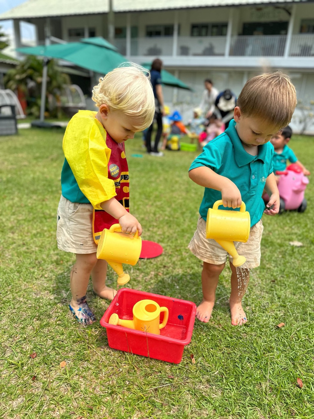 Two toddlers standing side by side on the grass at Little Forest, each tipping their own yellow watering can into the same red tray, both watching their own stream of water