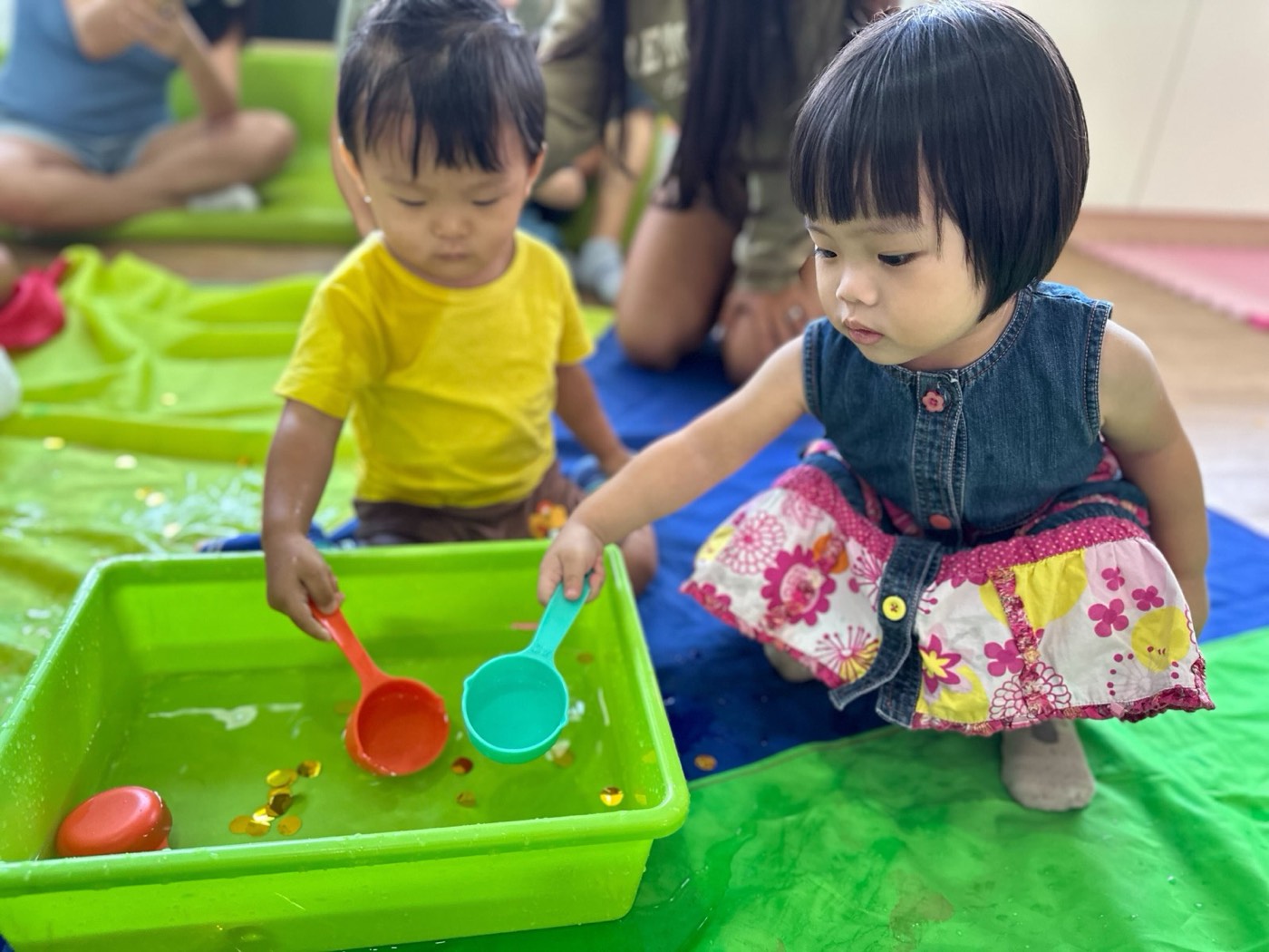 Two toddlers kneeling at the same green water tray at Little Forest in Singapore, each holding their own scoop, neither looking at the other