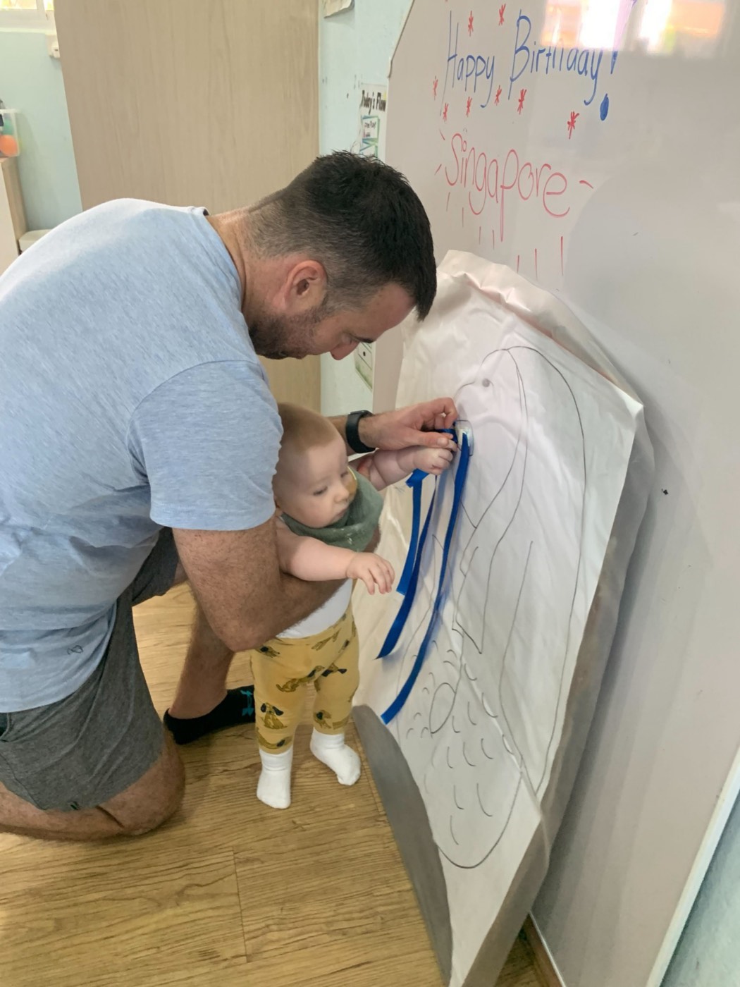 A father kneeling beside his baby at Little Forest, taping blue ribbon onto a large National Day mural together