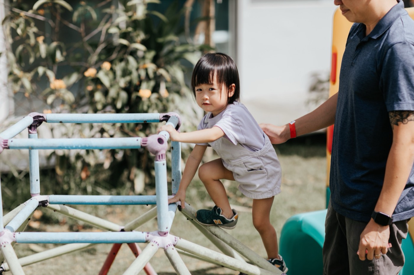A toddler climbing a pastel climbing frame outdoors at Little Forest, with her father's hand resting on her back