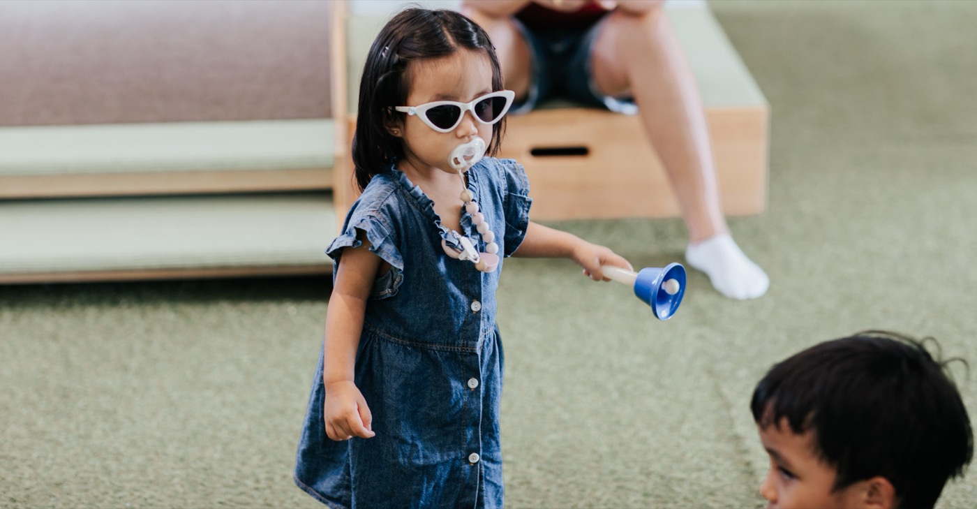 A toddler in a denim dress and white sunglasses standing in a Little Forest class in Singapore, ringing a blue handbell