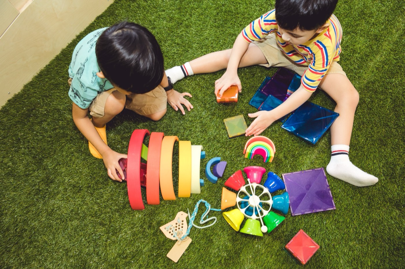 Two boys sitting on the turf a short distance apart at Little Forest, each with their own set of wooden and magnetic toys, both looking down at their own work