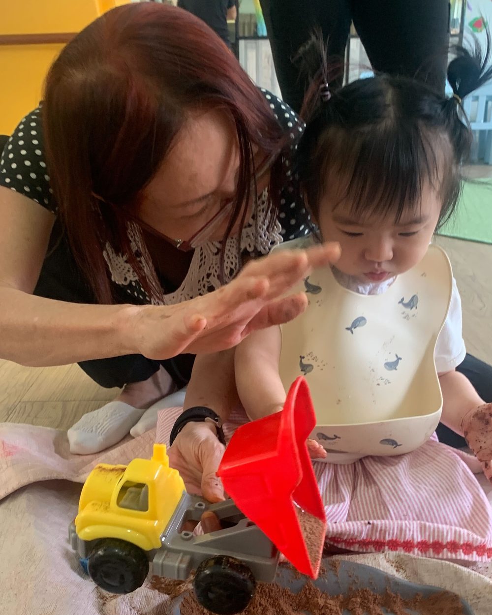 An adult in a polka dot top kneeling beside a toddler in a whale print bib, both with sand on their hands, the toddler tipping a red toy dump truck in a sand tray